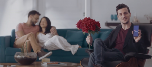 Imagem do ator segurando telefone com o app e o casal apaixonadão no fundo